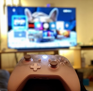 A gaming controller held in the foreground with a blurred television displaying a home screen interface in the background. The room has a cozy atmosphere with plants and a wooden shelf.