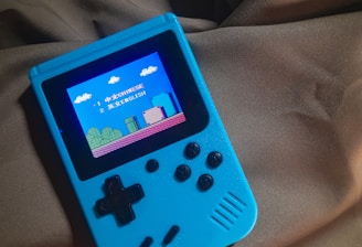 A handheld gaming device with a bright blue casing rests on a soft, dark fabric background. The screen displays a pixelated video game scene with options to select either Chinese or English language. The layout includes a classic D-pad and buttons, reminiscent of retro gaming consoles.